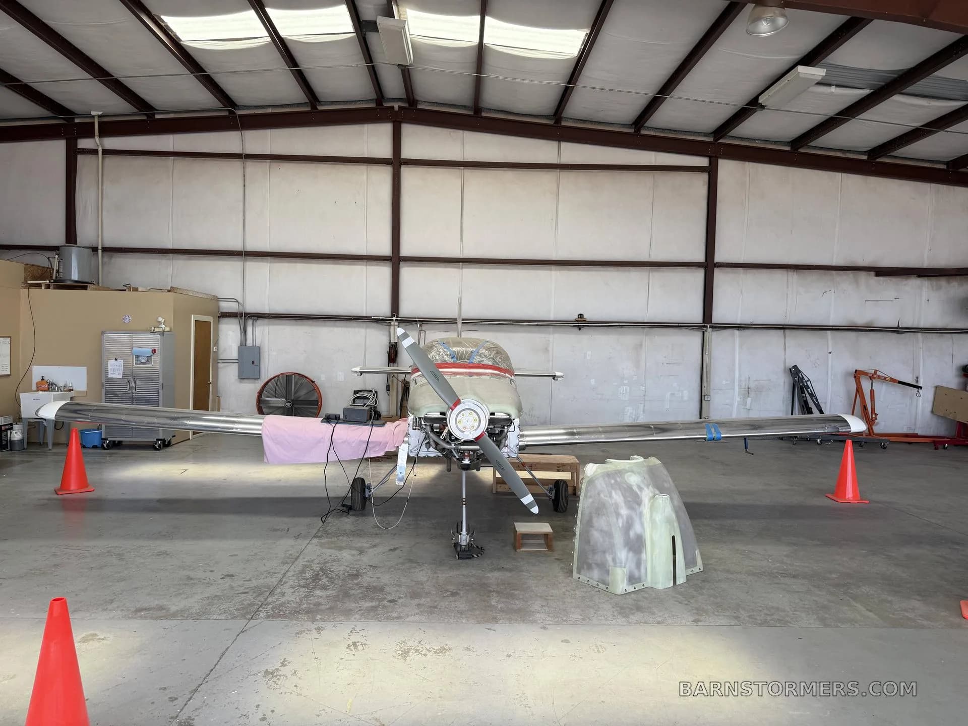 A front view of a partially assembled, unpainted metal aircraft with an exposed engine in a hangar.
