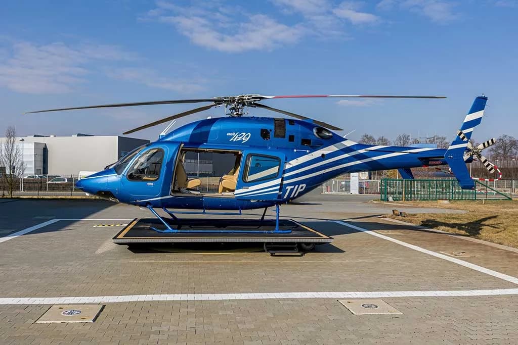 A side view of a blue helicopter with white stripes, registration OM-TIP, on a helipad with its passenger door open.