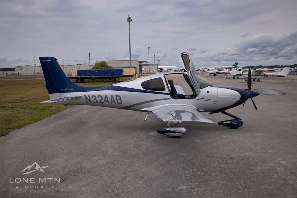A white and blue Cirrus SR22T aircraft with open doors parked on an airfield, viewed from the front-right.
