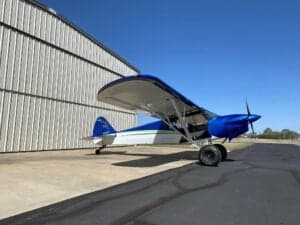 A blue and white Cubcrafters aircraft is parked outside a hangar on a sunny day.
