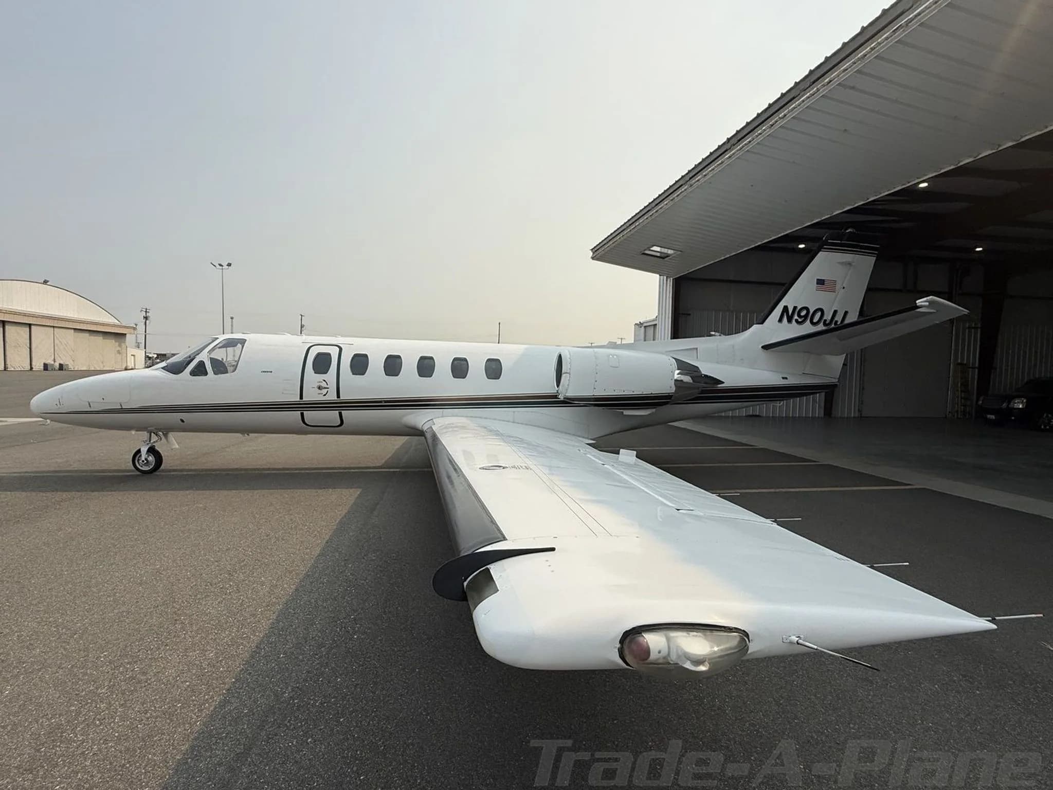 A white Cessna Citation jet, N90JJ, with black and gold stripes, seen from the rear-right wing, parked near a hangar.