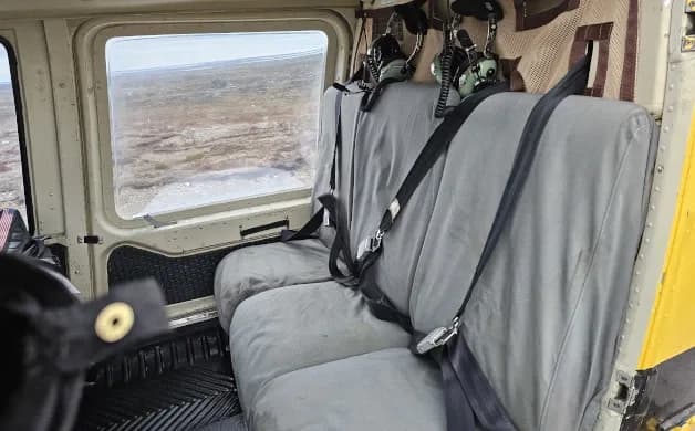 The interior of a helicopter cabin shows two grey passenger seats with seatbelts and a view of a barren landscape through the window.