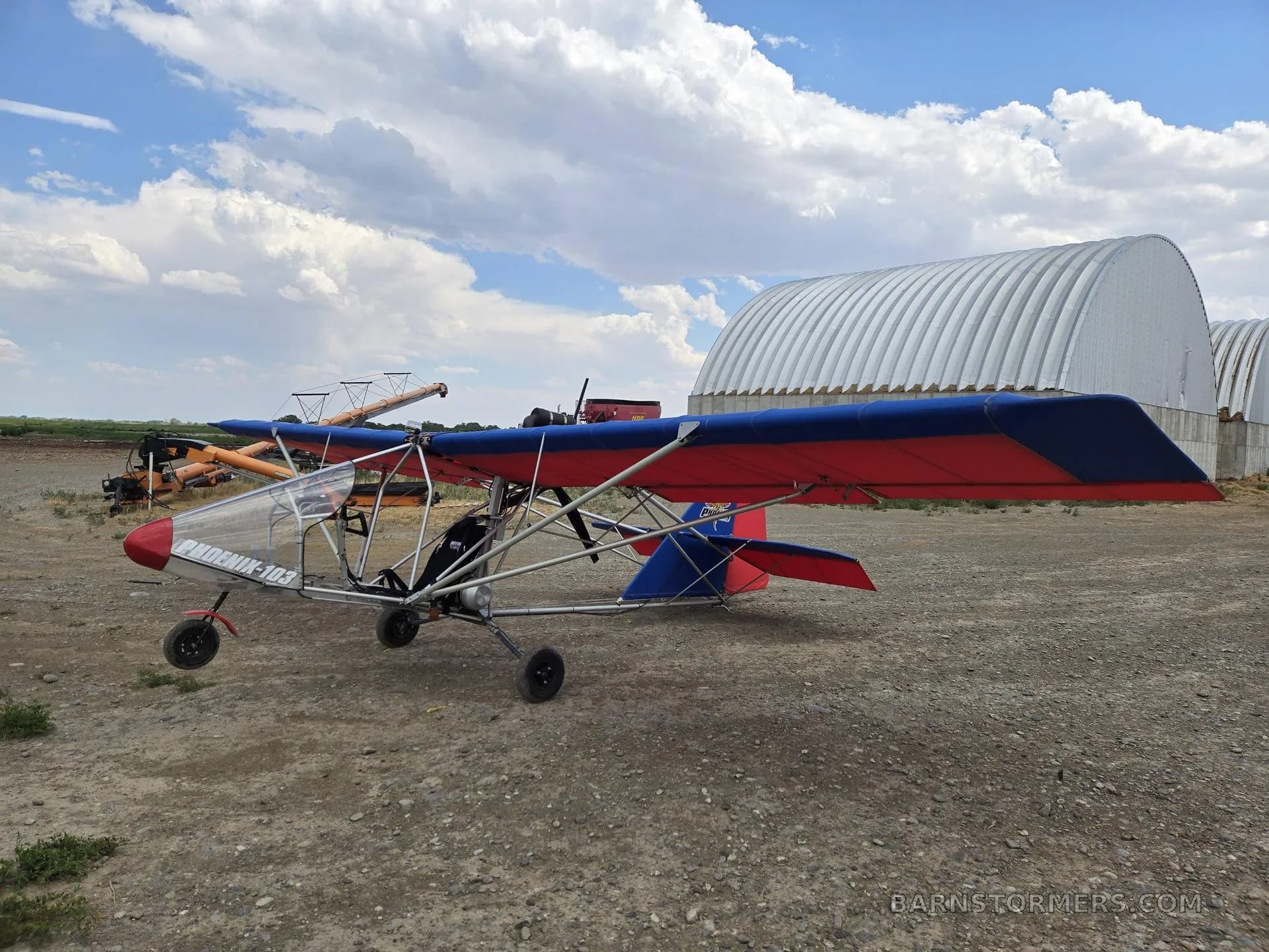 A red and blue ultralight aircraft with a clear canopy, parked on a dirt field in front of large white hangars.