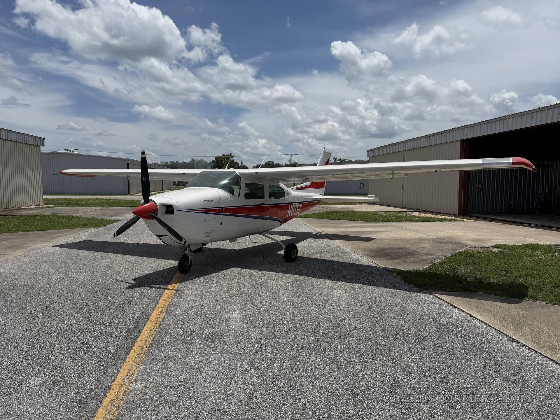 A white and red Cessna 210 Centurion aircraft is parked on an asphalt ramp between hangars under a partly cloudy sky.