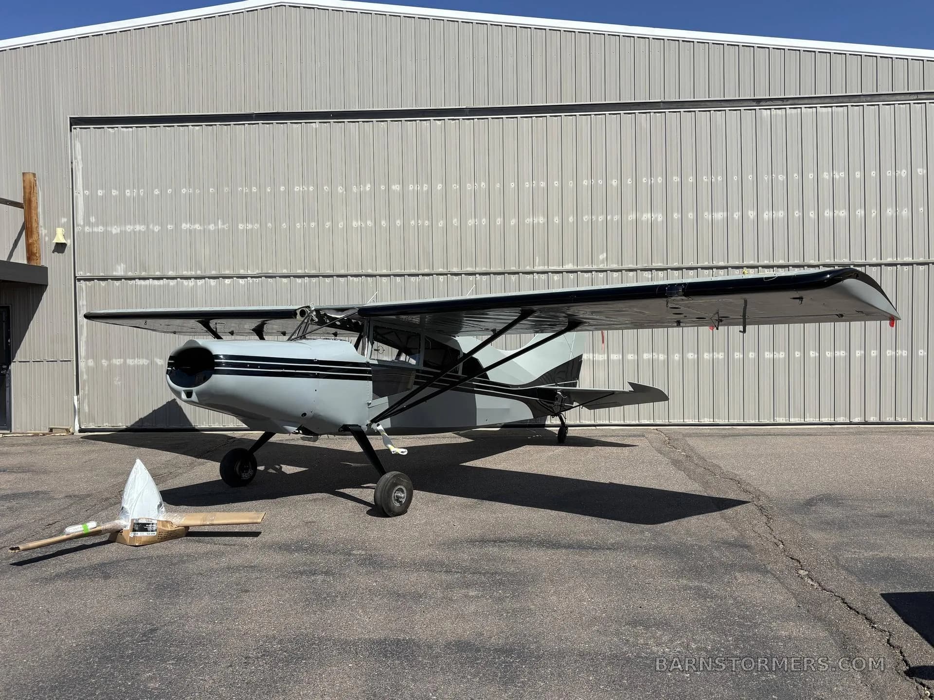 A grey Maule M-7 aircraft with black stripes parked in front of a large hangar door.