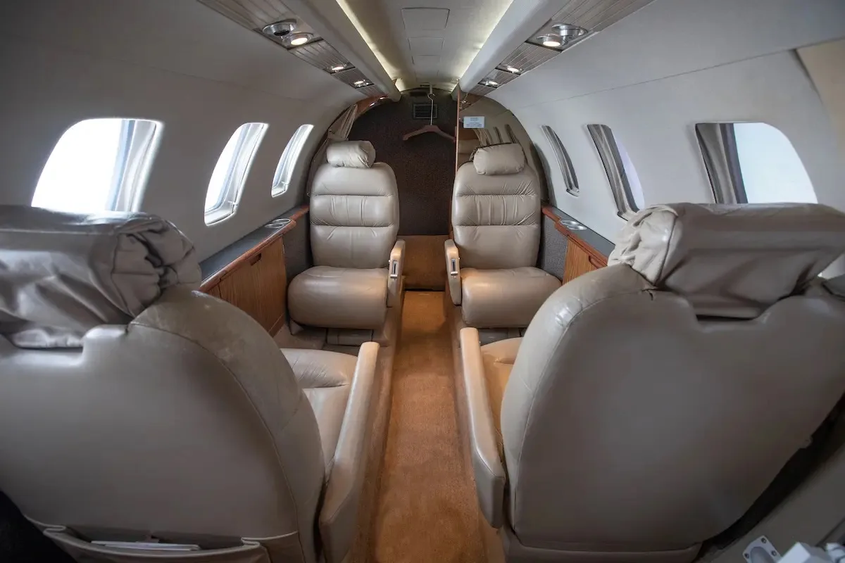 The aircraft cabin interior features four tan leather seats facing each other, with wood trim and windows.