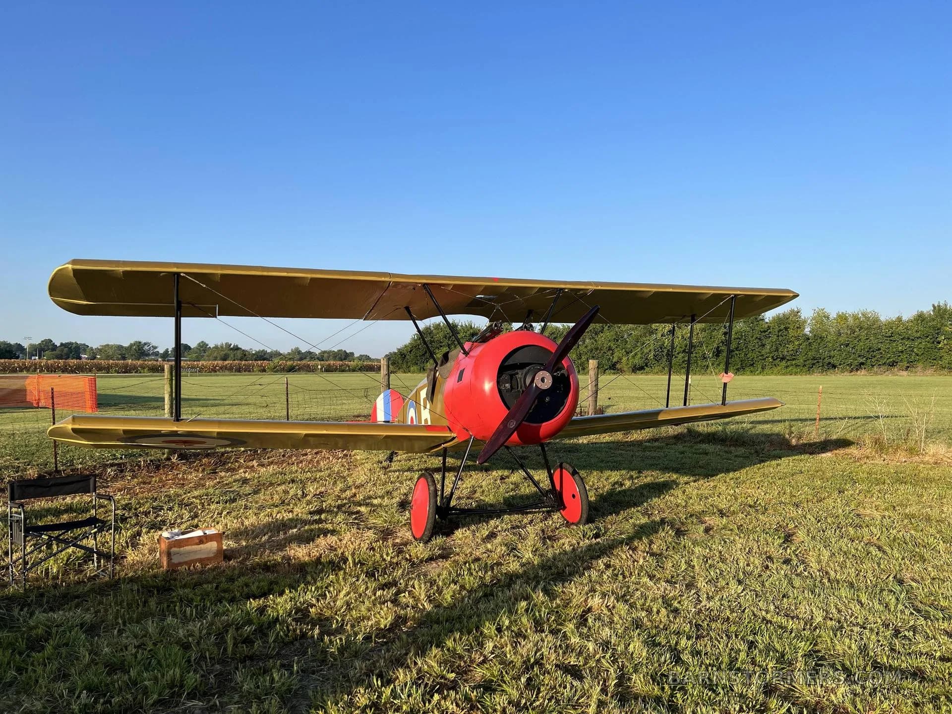 A tan and red biplane with a red propeller is parked on a grassy field under a clear blue sky.