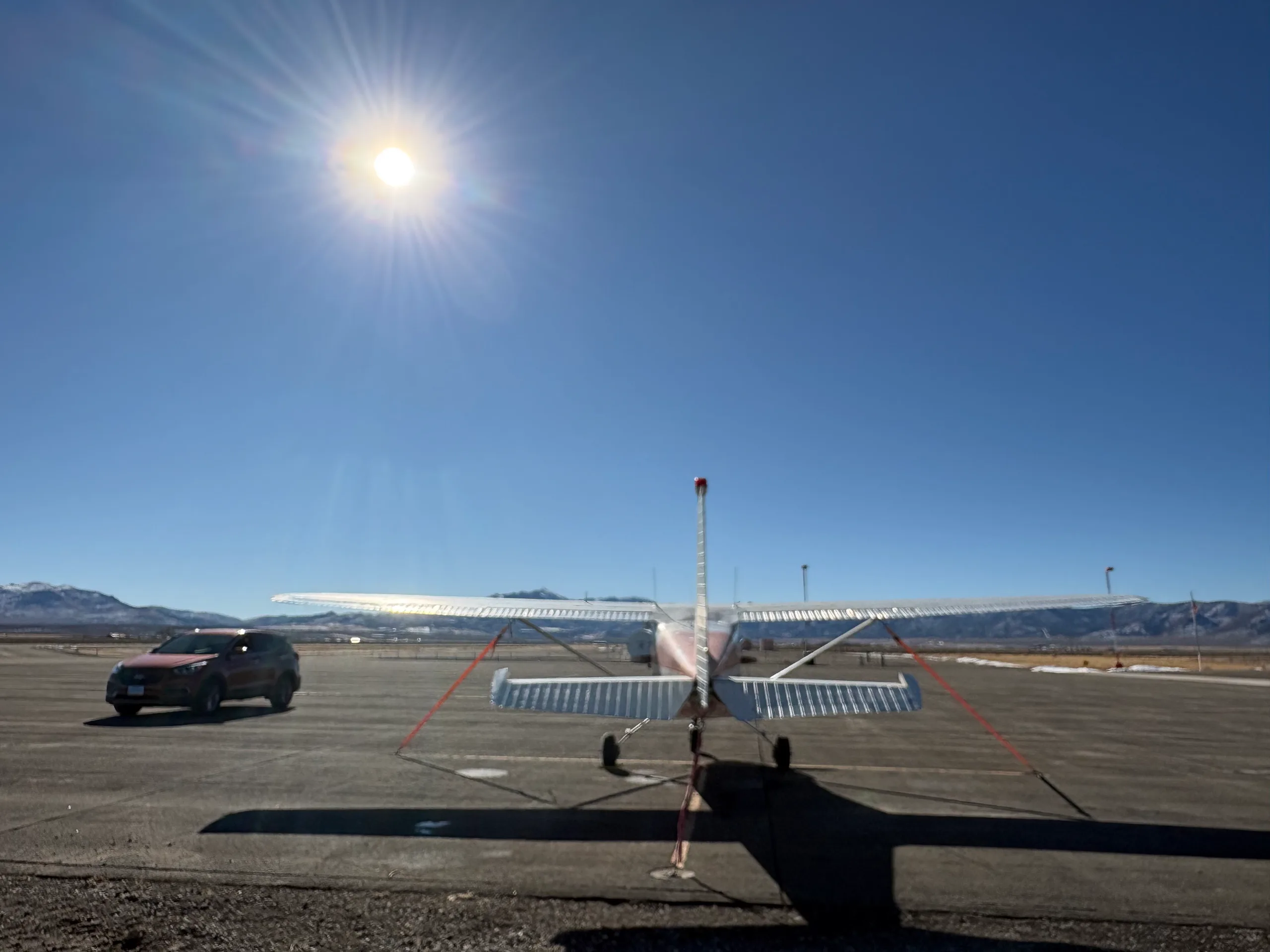 A red and white single-engine high-wing aircraft, N5799A, viewed from behind on an airfield with a red car and bright sun.