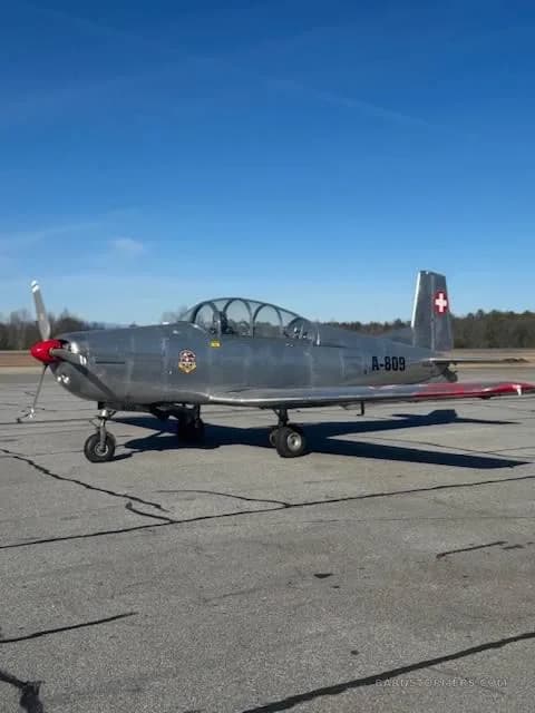 A silver Pilatus P-3 trainer aircraft with a red propeller spinner and Swiss flag on the tail, parked on an asphalt runway under a clear blue sky.