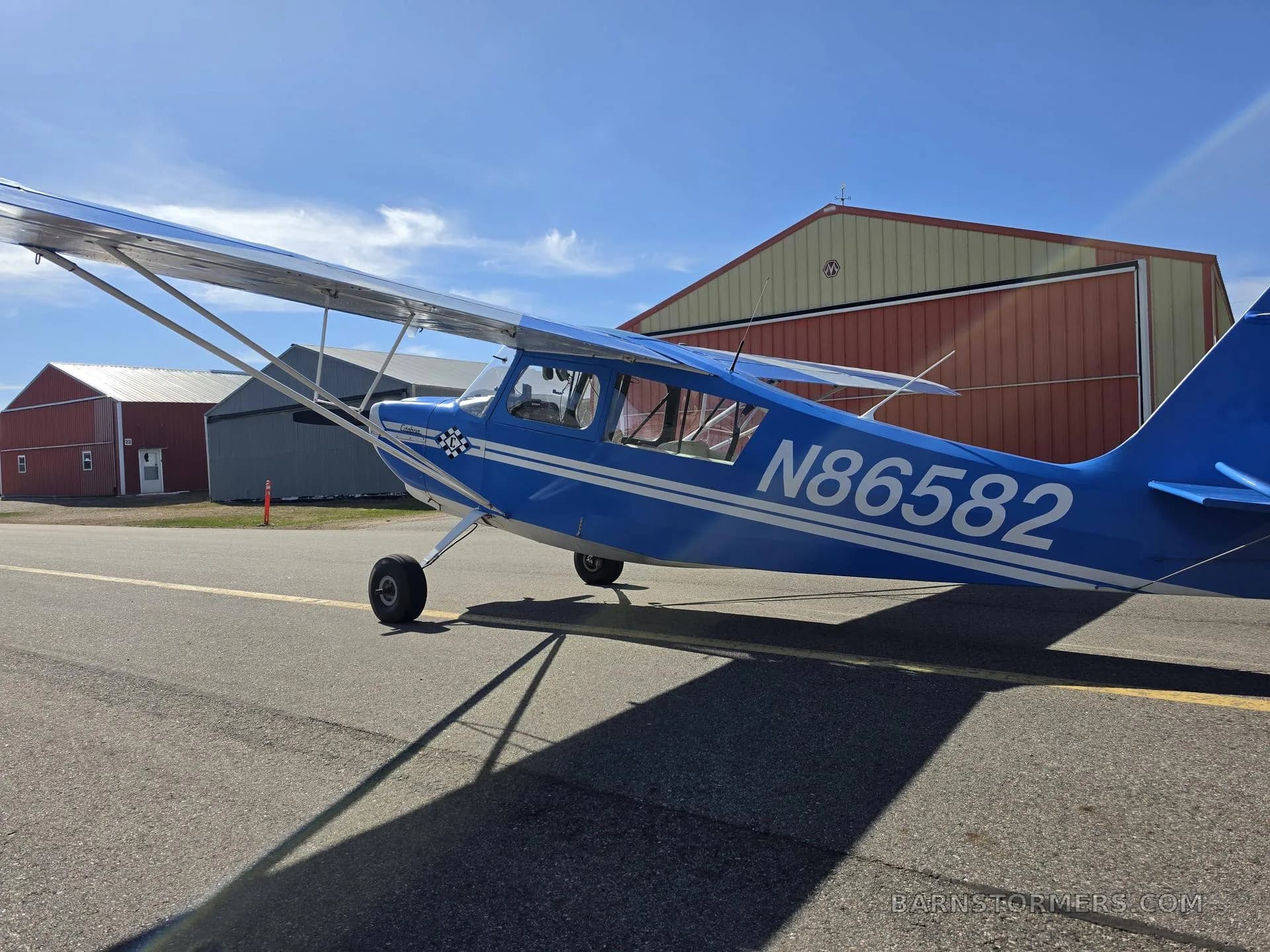 A blue and white single-engine propeller aircraft, tail number N86582, parked on an asphalt ramp with red and grey hangars in the background.