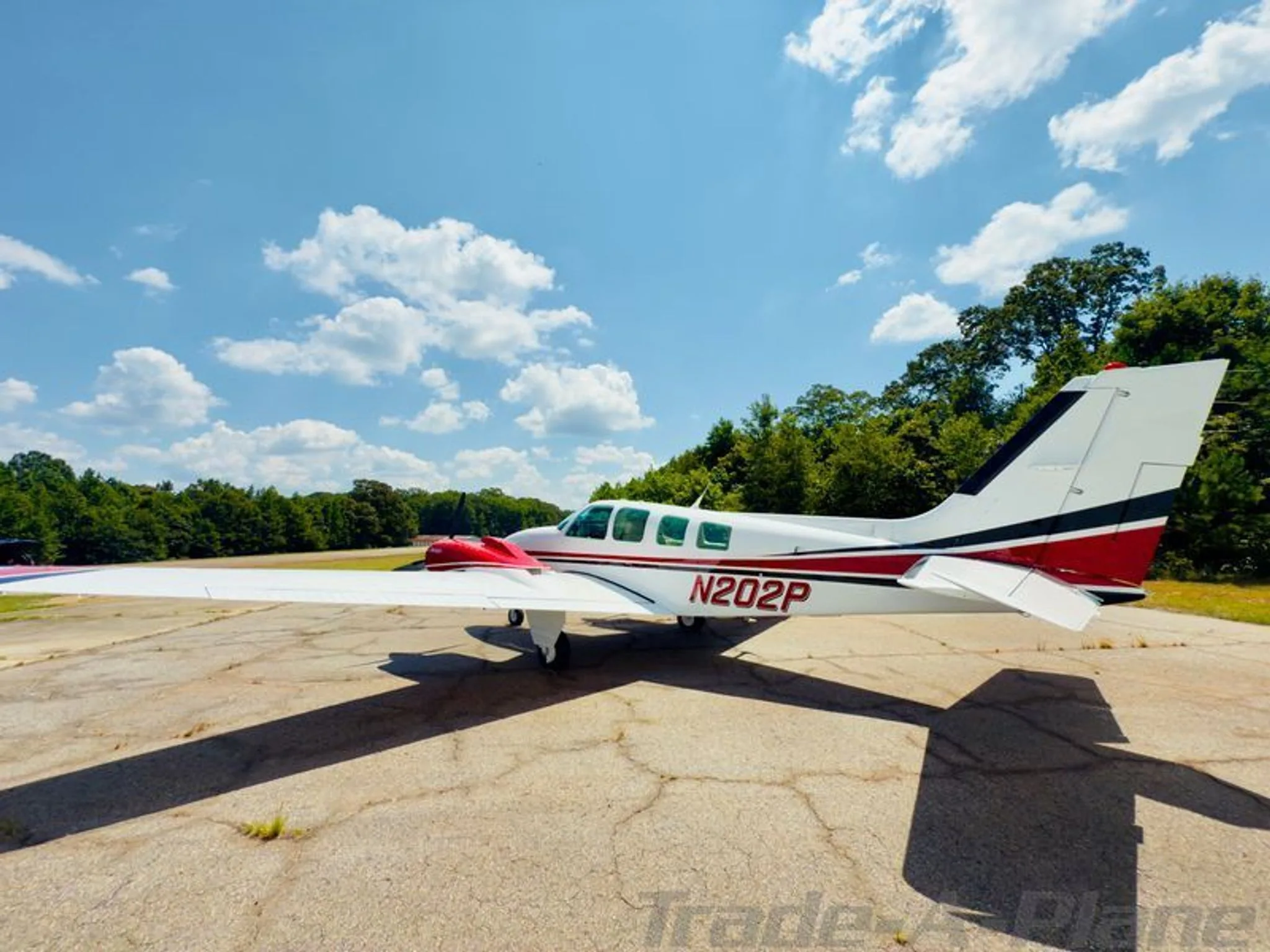 Side view of the white Beechcraft Baron N202P with red and black stripes, parked on a cracked asphalt surface under a blue sky.