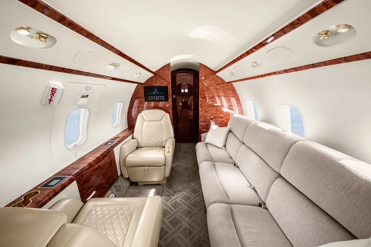 A business jet cabin interior featuring a long grey sofa and a single cream leather seat with polished wood trim.