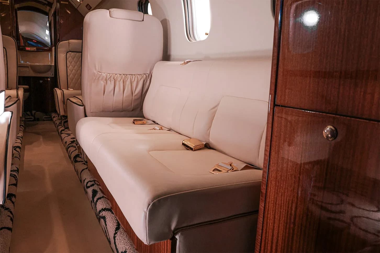 A cream leather couch-style seat with gold seatbelts in the private jet's cabin, next to dark wood cabinetry.