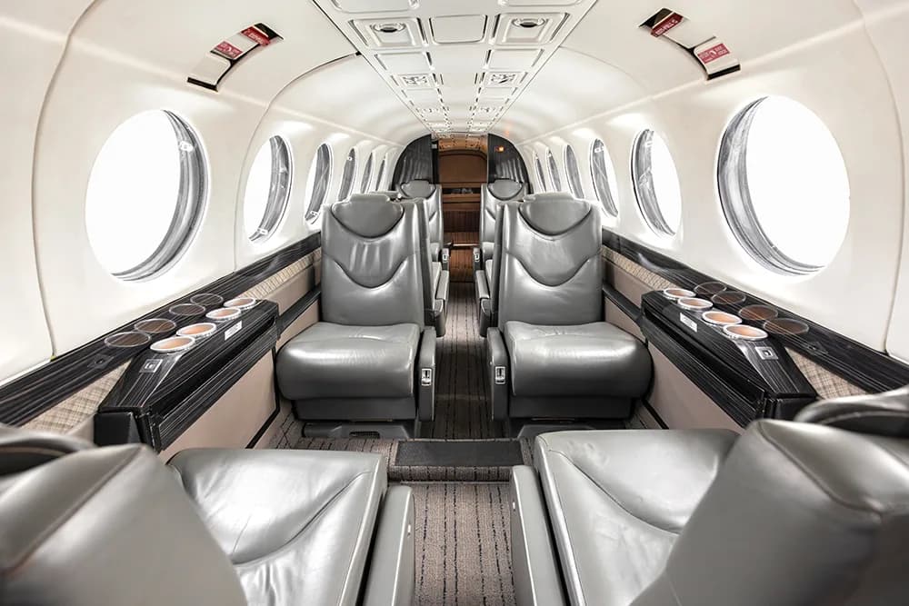 A view down the aircraft cabin aisle with grey leather seats, open tables with cup holders, and large circular windows.