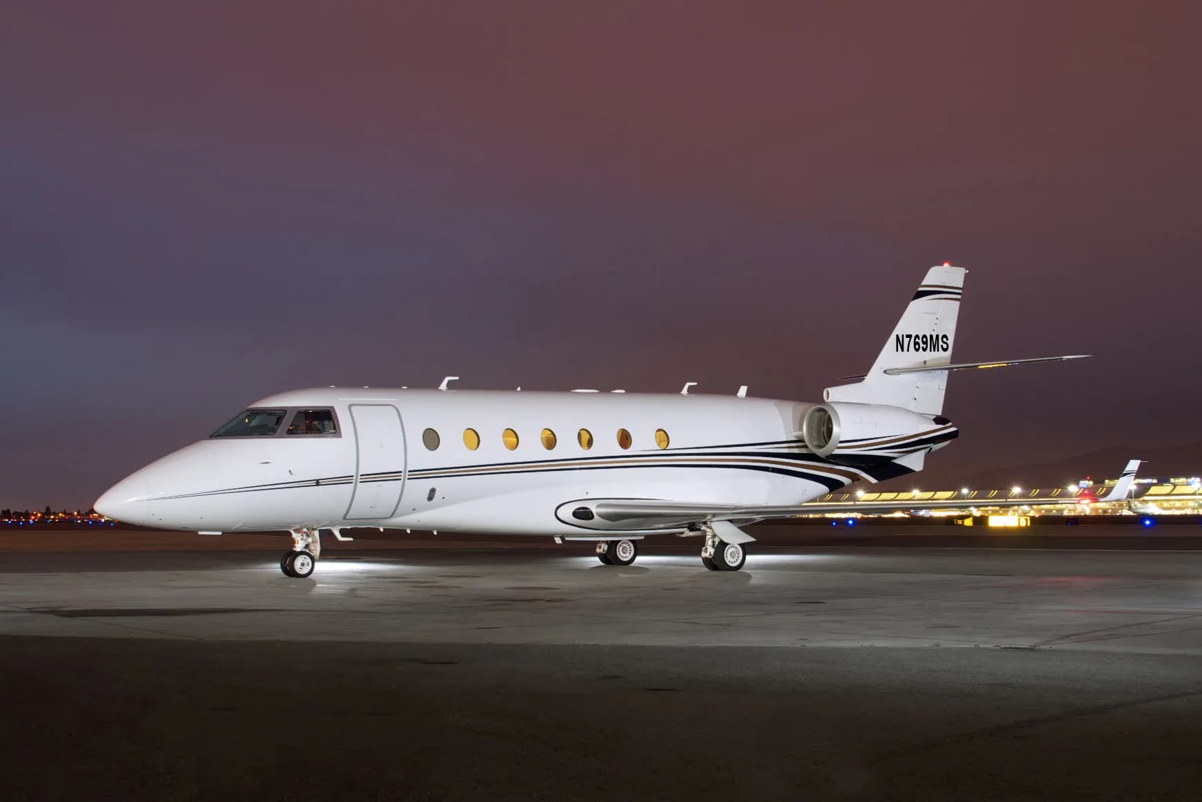 A white business jet with black and gold stripes on the fuselage and tail, parked on a tarmac at night with airport lights in the background.