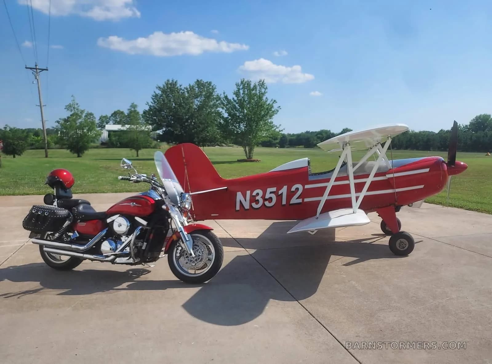 A red biplane with white wings and N3512 on the fuselage, parked next to a red motorcycle on a concrete pad.