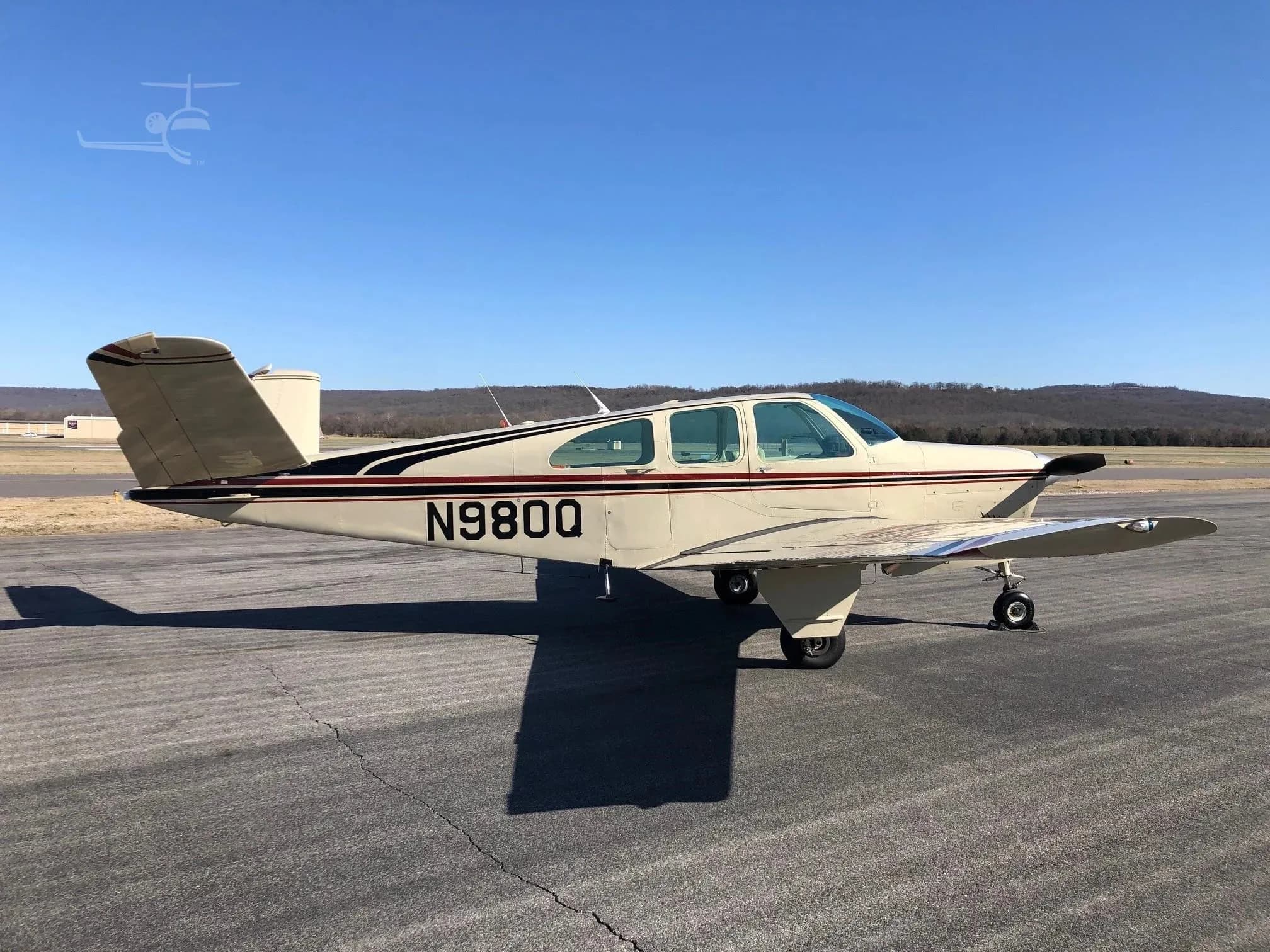 A side view of a beige Beechcraft Bonanza N980Q with red and black stripes parked on an asphalt runway.