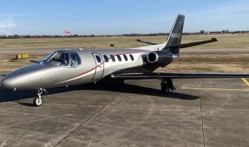 A silver Cessna Citation jet with a red stripe and dark tail parked on an airport tarmac under a clear sky.