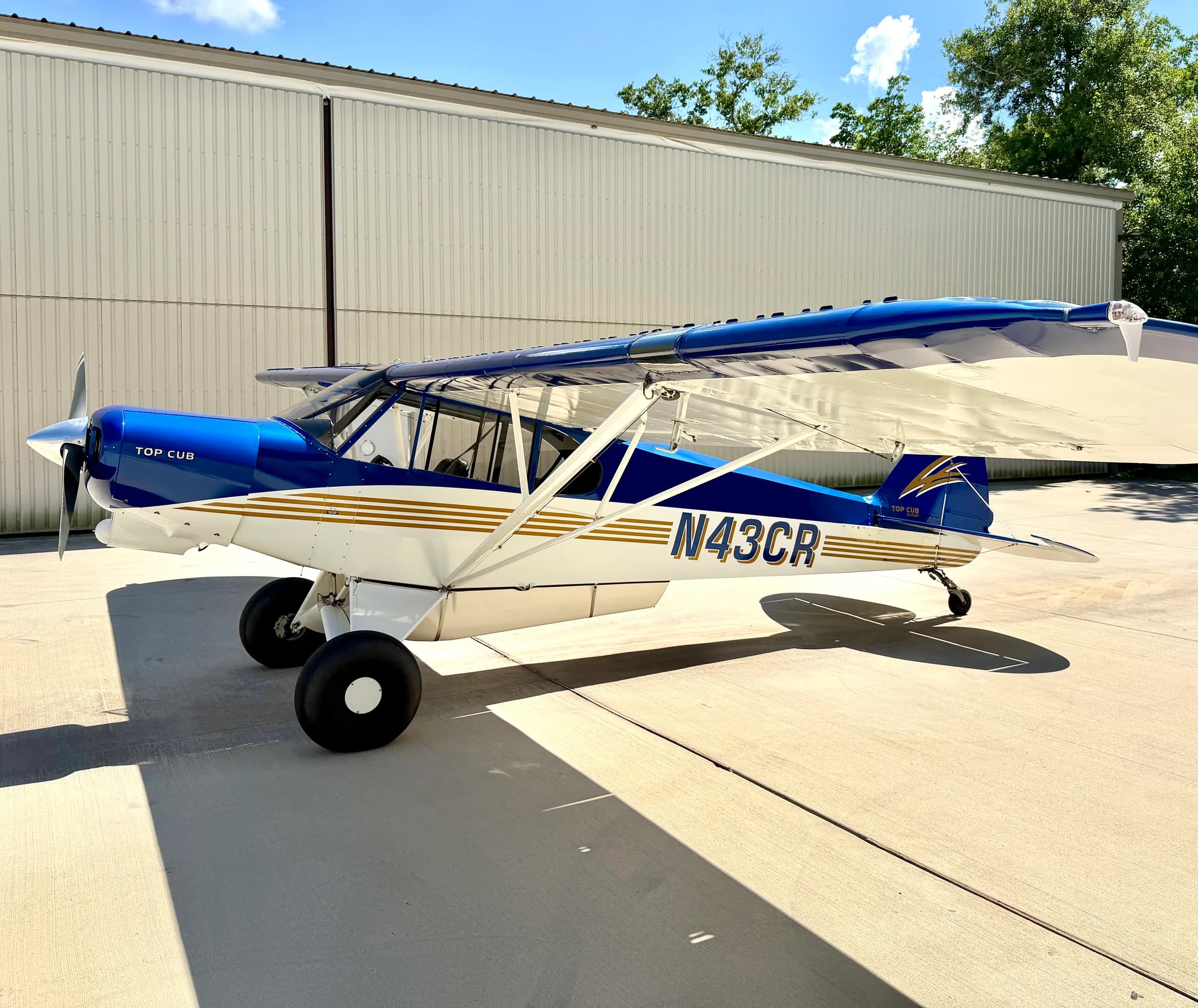 A blue and white Top Cub aircraft with gold stripes is parked on a tarmac in front of a white hangar under a blue sky.