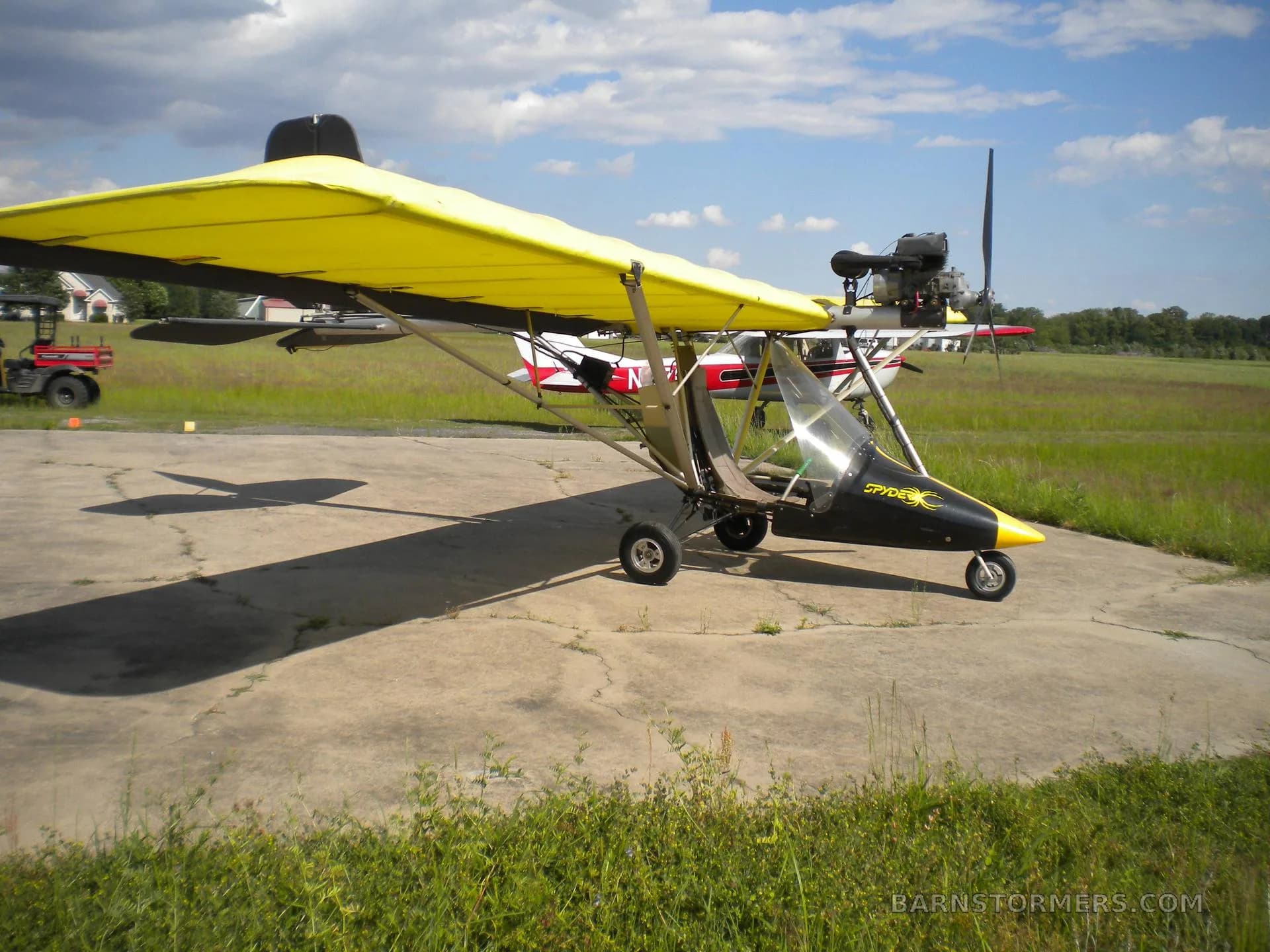 A yellow-winged ultralight aircraft with a black nose cone and 'Spyder' logo, parked on a concrete pad at an airfield.