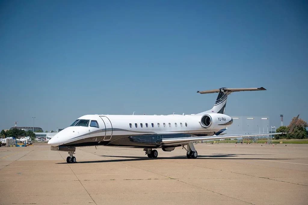 A white and black Embraer Legacy 600 business jet with registration YU-PAA parked on a tarmac under a clear blue sky.
