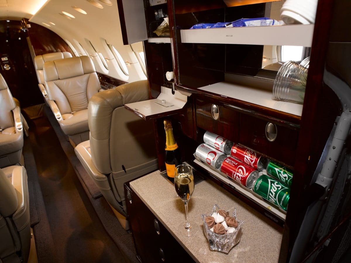 The private jet's galley area features a mini-fridge stocked with drinks and a counter with champagne.