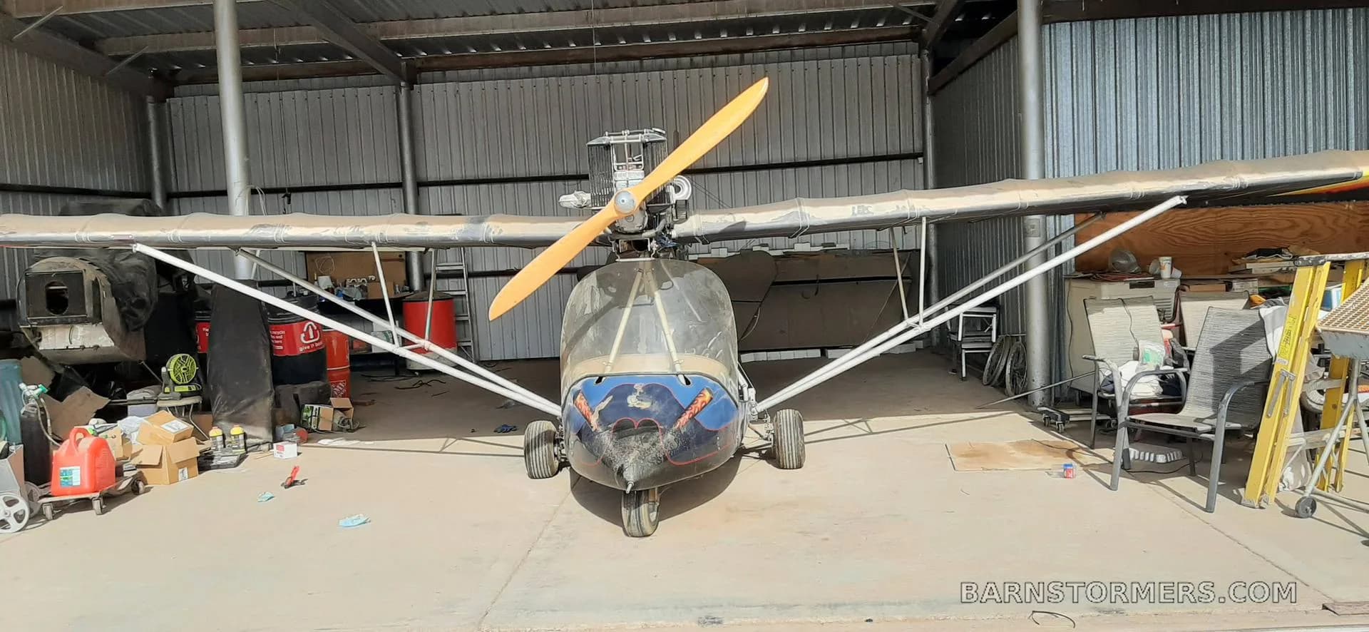 A front view of a blue and black ultralight aircraft with an exposed engine and propeller, parked inside a dusty hangar.