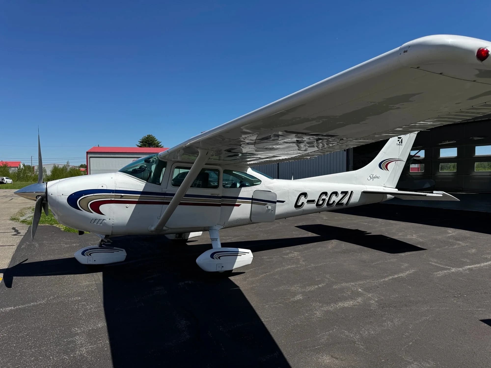 A white Cessna Skylane (C-GCZI) with blue, red, and gold stripes is parked on an asphalt ramp under a clear blue sky, with a hangar in the background.