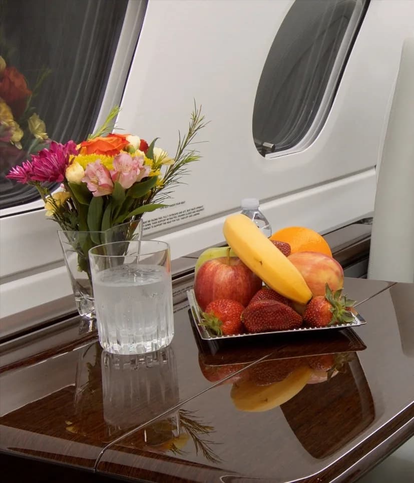 A close-up of a polished wooden table inside the aircraft cabin, displaying a fruit platter, a glass of water, and a bouquet of flowers.