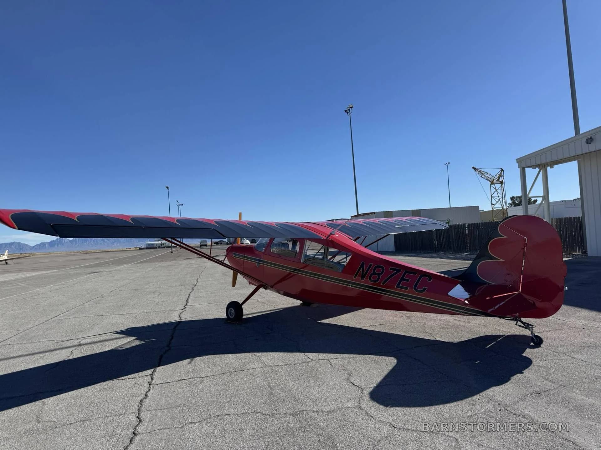 A red high-wing monoplane with black and grey wing accents and registration N87EC is parked on an airfield with mountains in the background.