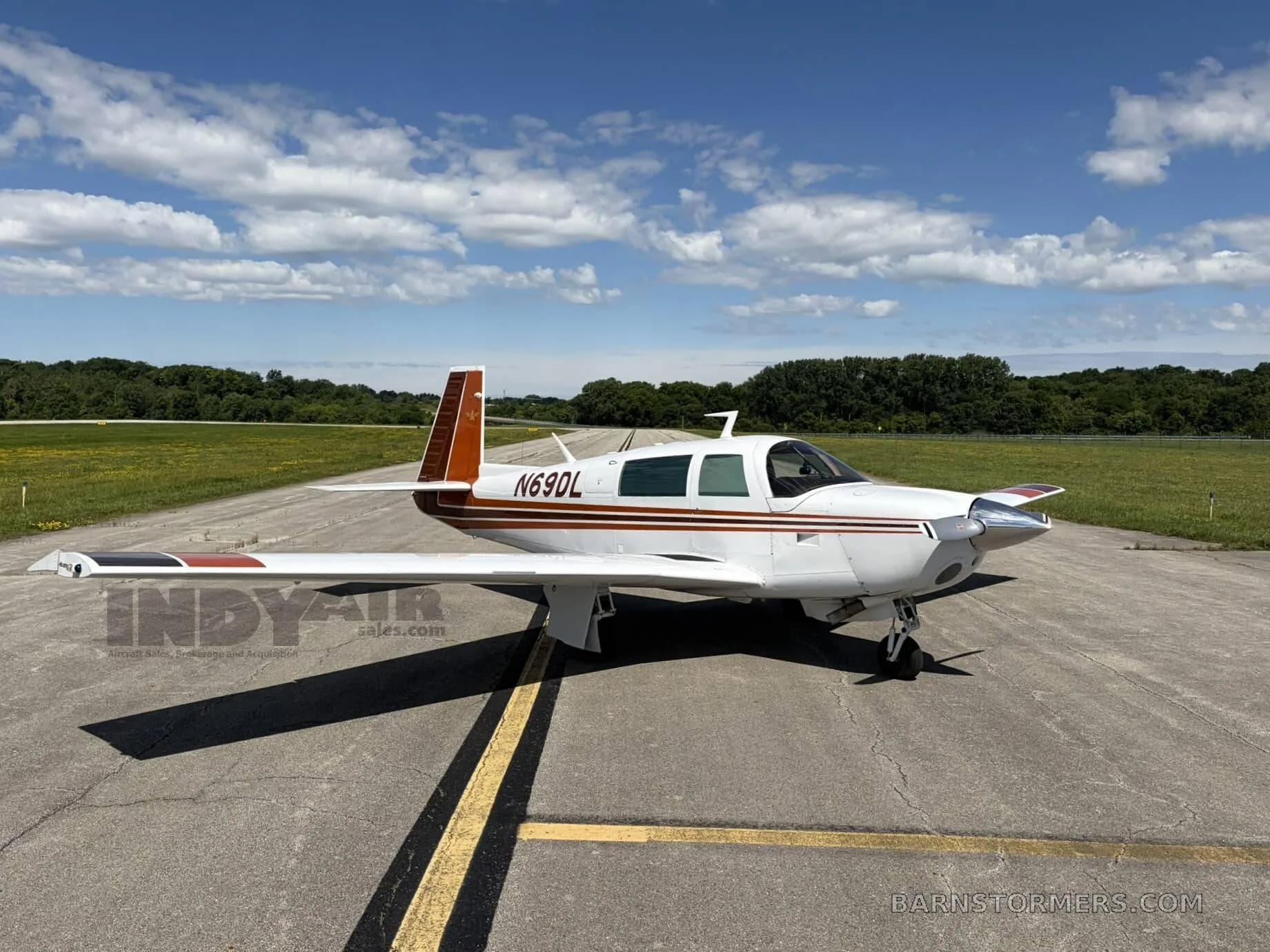 A white and brown Mooney M20J aircraft (N69DL) is parked on an asphalt runway under a partly cloudy sky, viewed from the front-right side.