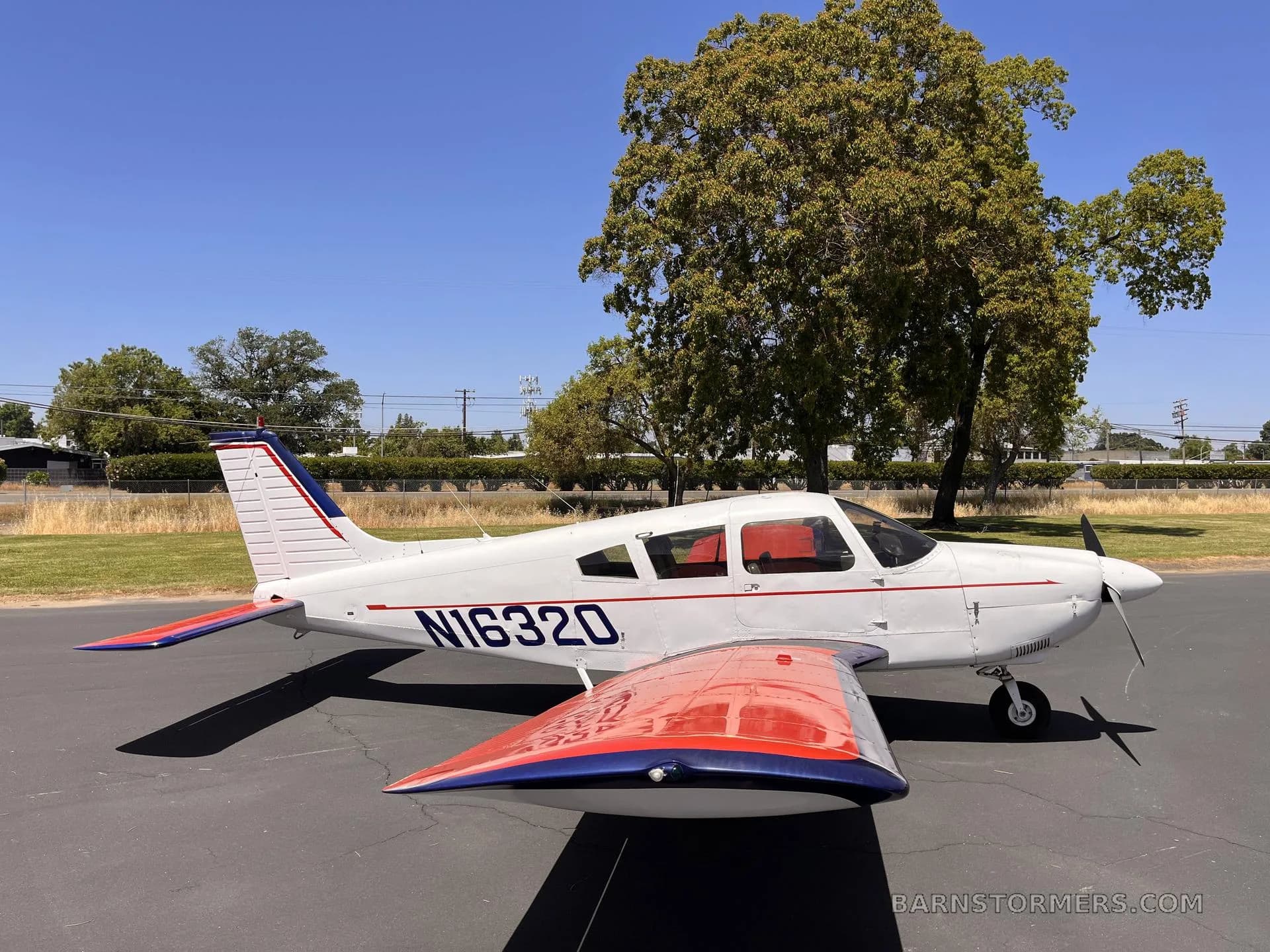 A white Piper Cherokee aircraft with red and blue accents on the wings and tail, parked on an asphalt ramp with trees in the background.