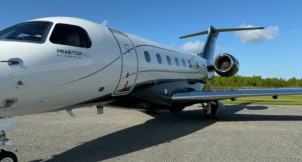 A close-up side view of a white and dark grey Embraer Praetor 600 business jet on an airfield, showing the 'PRAETOR 600' branding.