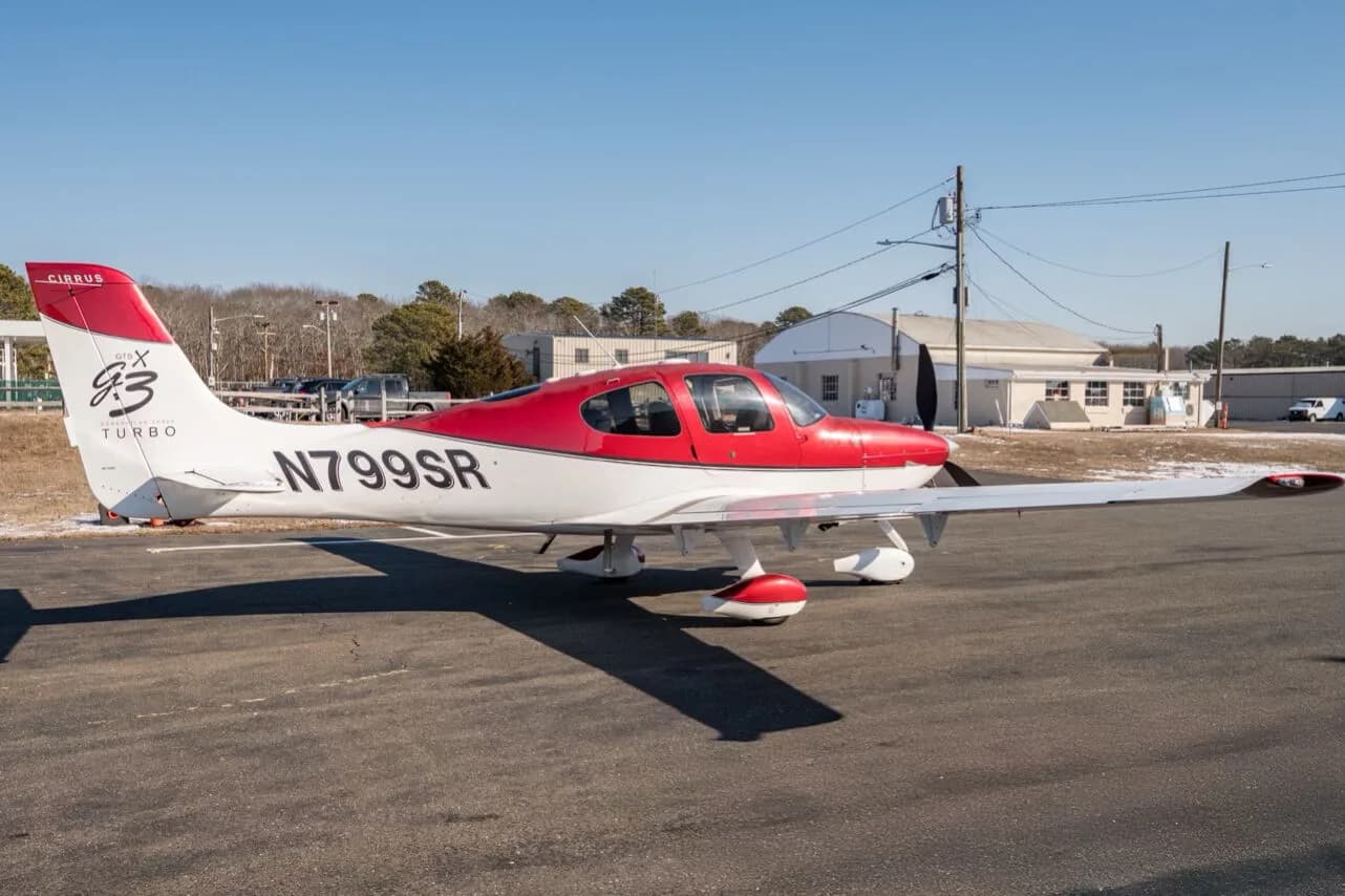 A red and white Cirrus SR22T aircraft, N799SR, parked on an tarmac with buildings and trees in the background.