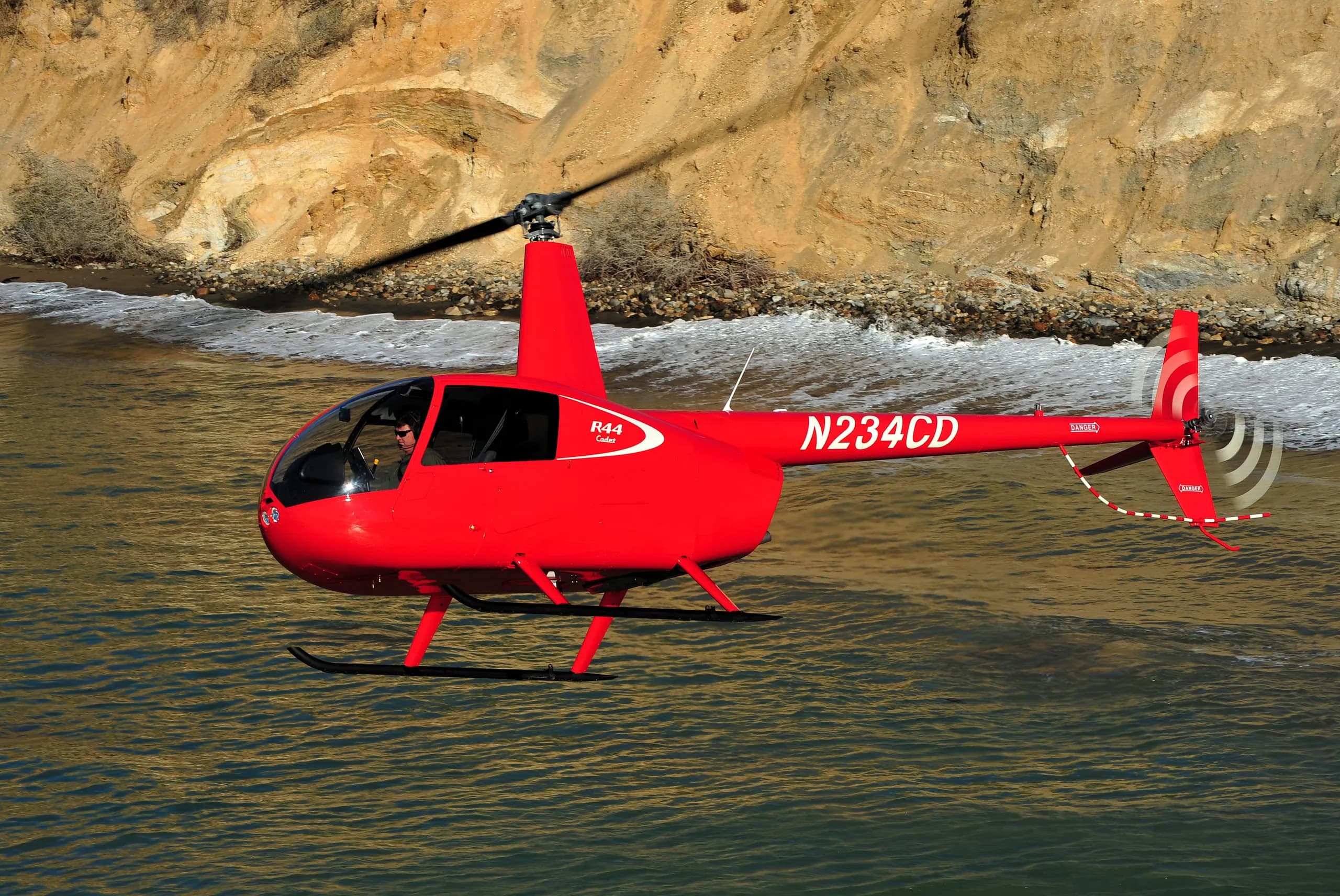 A red Robinson R44 helicopter flying over the ocean.