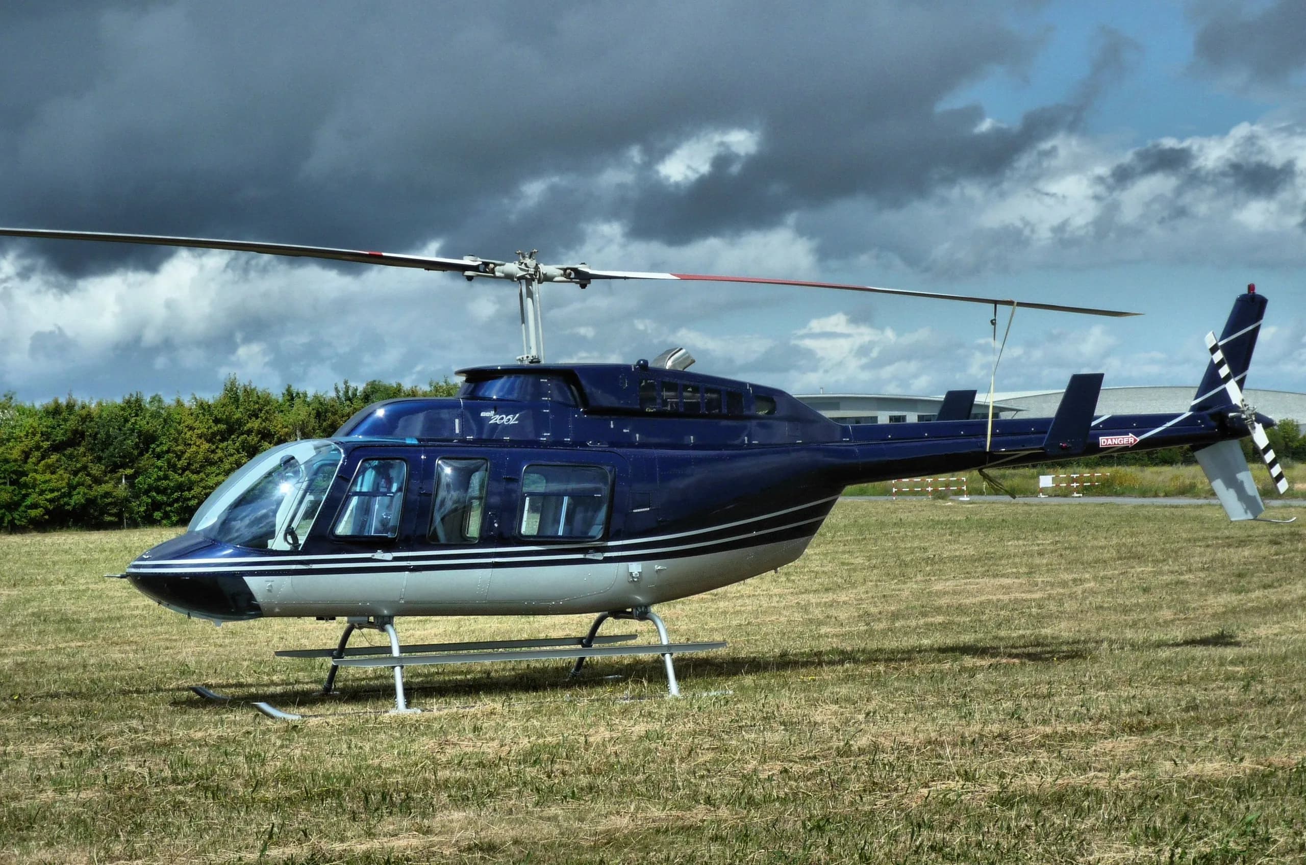 A blue and white Bell 206 JetRanger helicopter parked on a grassy field under a cloudy sky.