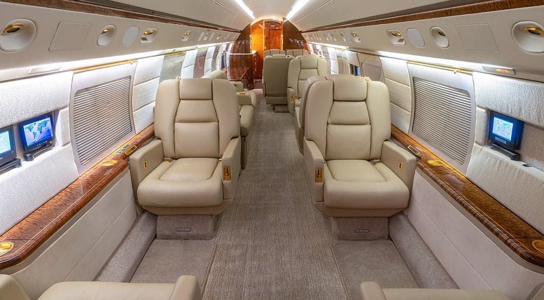 The spacious cabin interior of a private jet, featuring beige leather seats, wood accents, and individual entertainment screens.