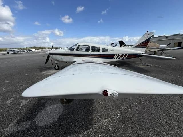 A white Piper Cherokee aircraft N7JJ with blue and red stripes, parked on an airport tarmac with other aircraft in the background.
