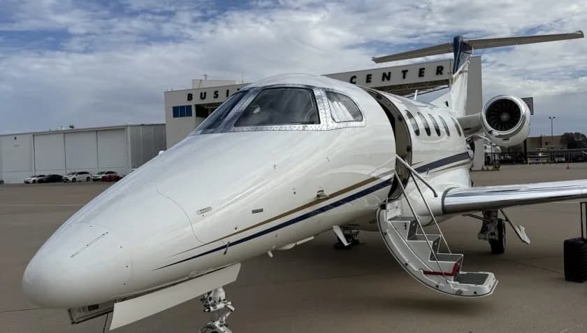 A close-up front view of a white Embraer Phenom 300 private jet with its passenger door open and stairs extended, parked in front of a business center.