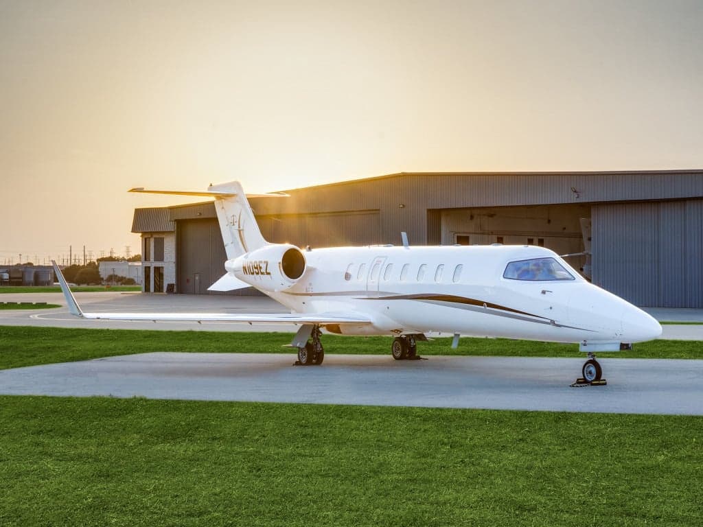 A white business jet (N109EZ) with gold and black accents is parked on a tarmac at sunset, with hangars in the background.