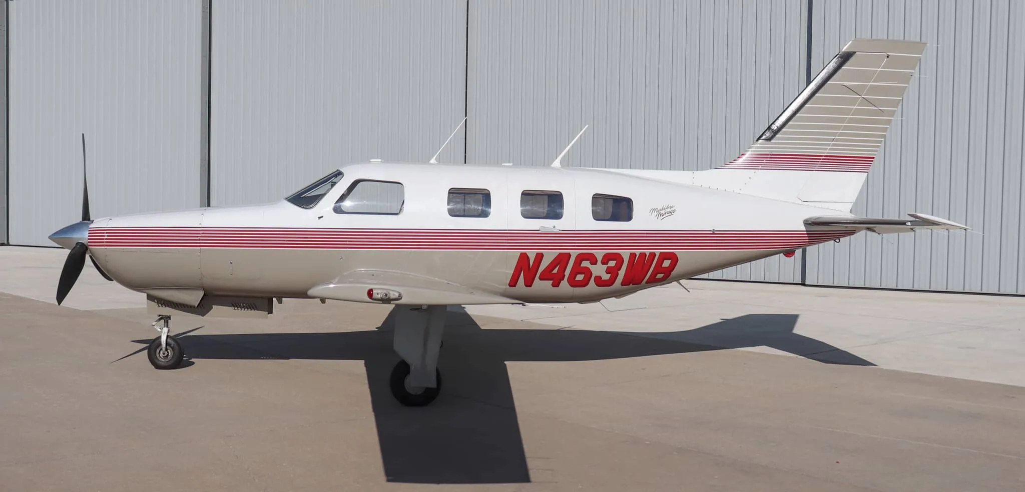 A full side profile of the Piper Malibu Mirage N463WB, featuring its white and tan livery with red stripes, parked in front of a hangar.