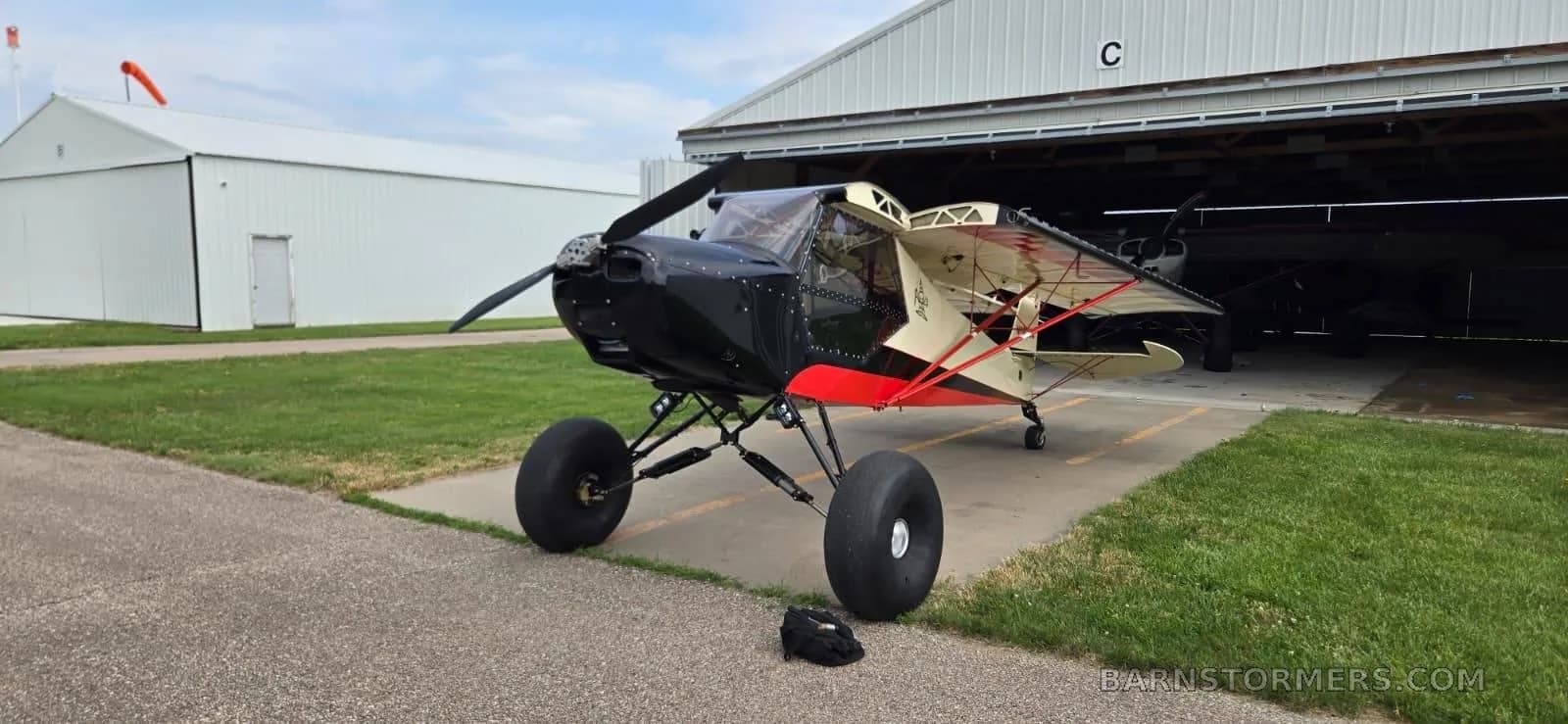 A black, red, and cream bush plane with large tires is parked on an asphalt ramp in front of an open hangar.