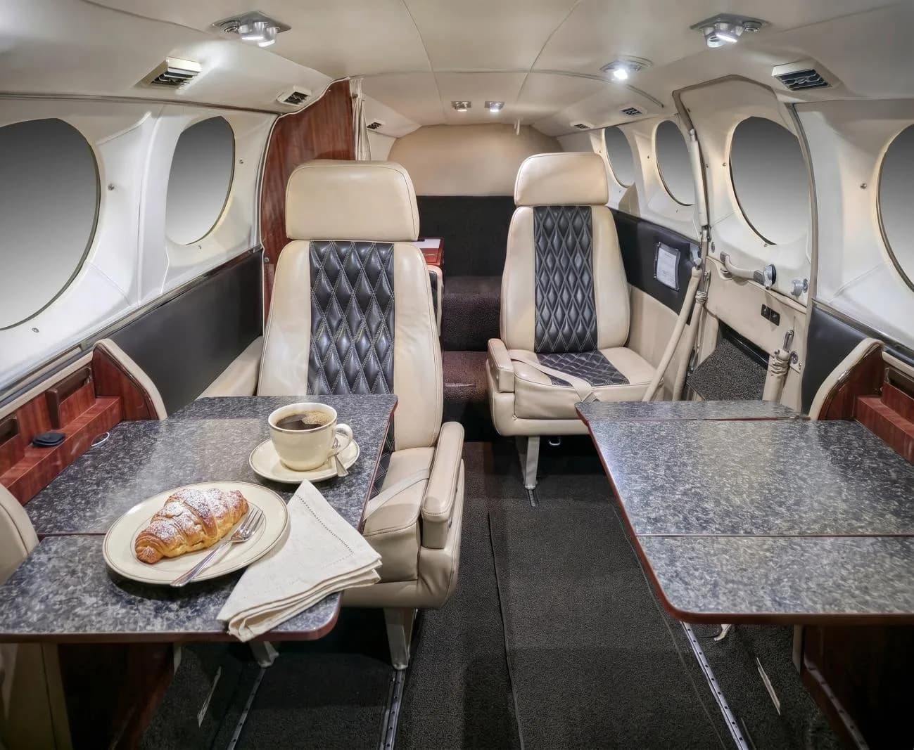 The aircraft cabin interior with beige and black quilted seats, two tables, and a croissant with coffee.