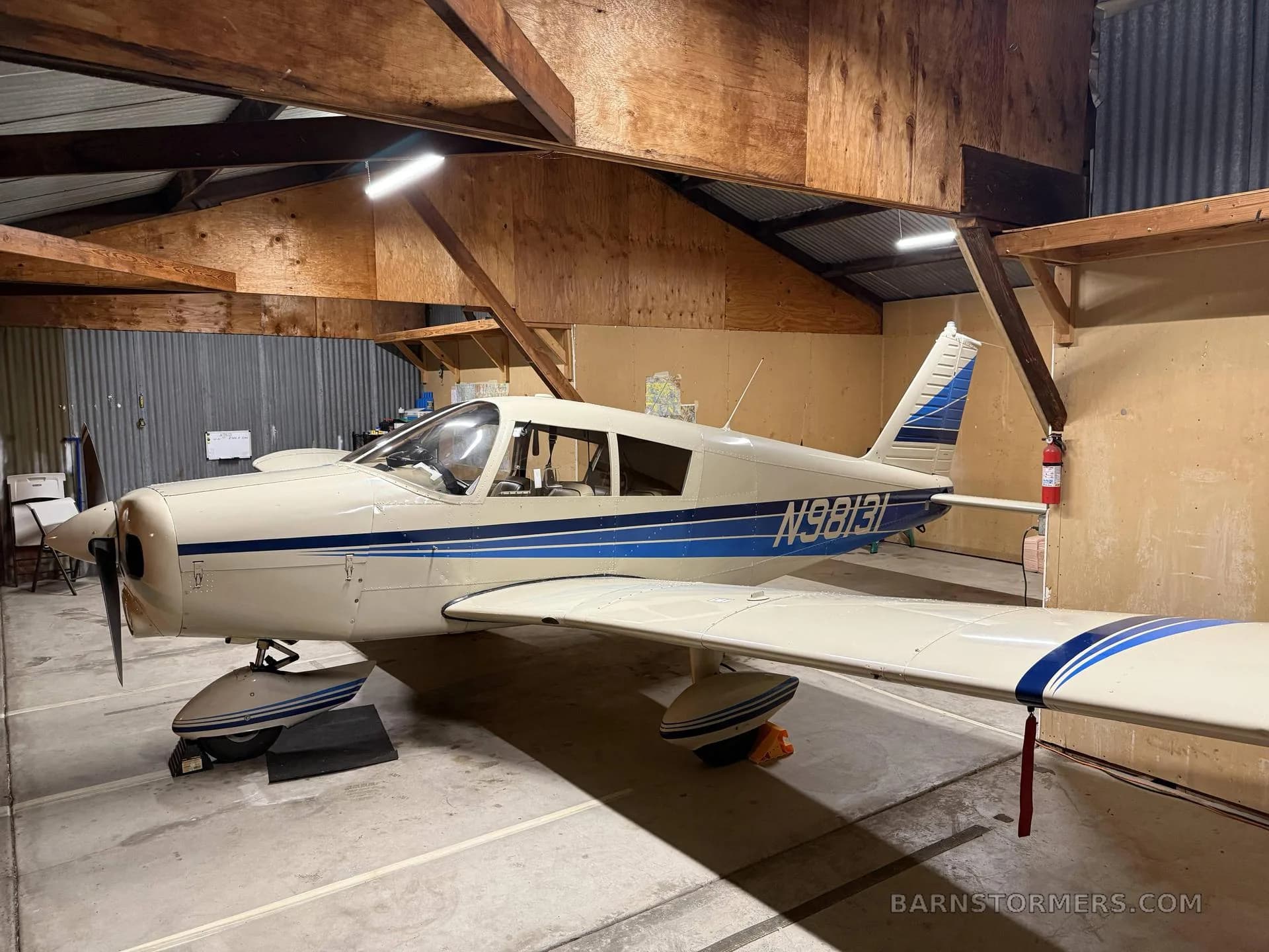 A white and blue single-engine propeller aircraft (N9813I) is parked inside a rustic hangar.