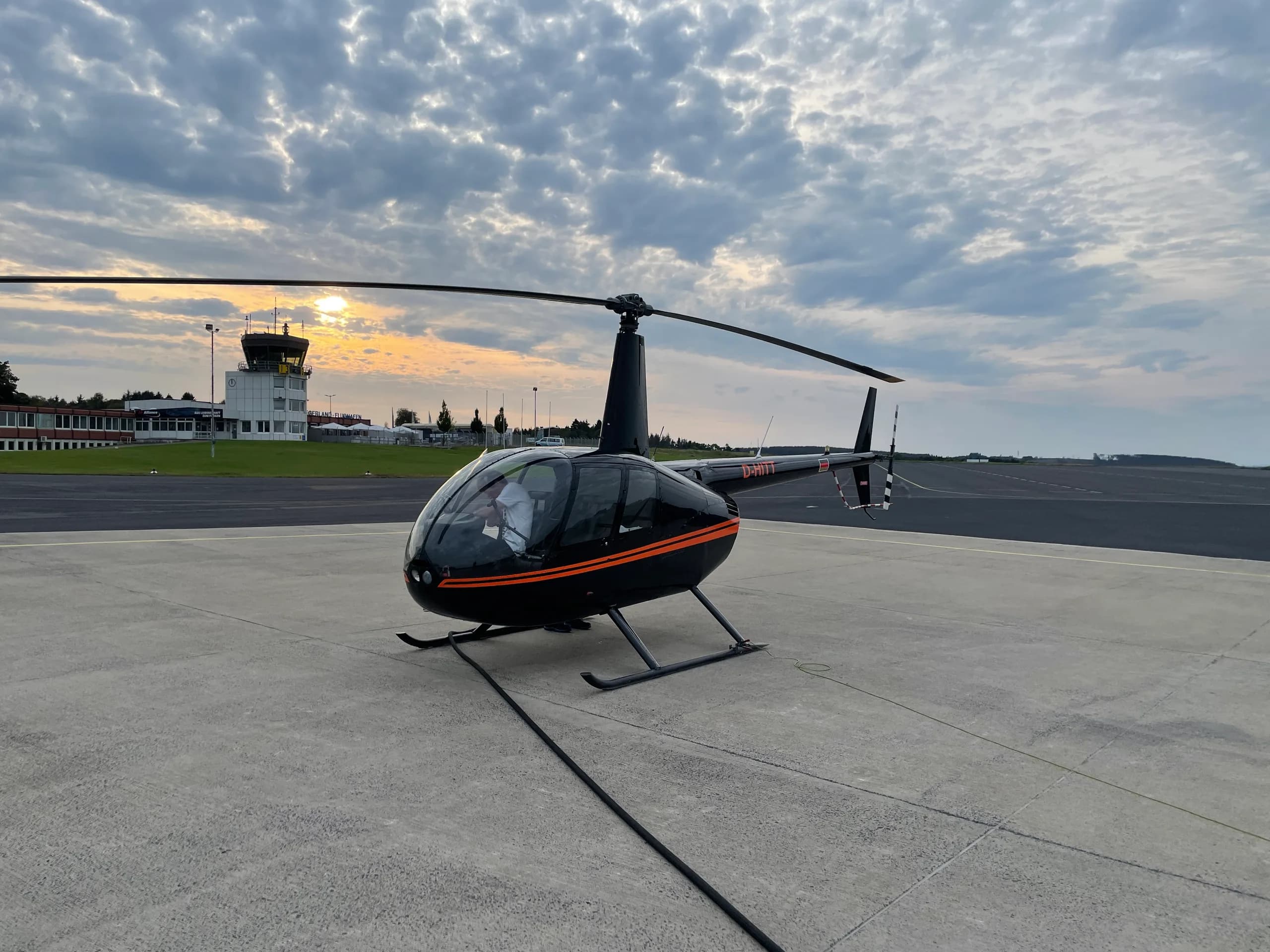 A black Robinson R44 helicopter with orange stripes is parked on an airfield at sunset, with a control tower in the background.