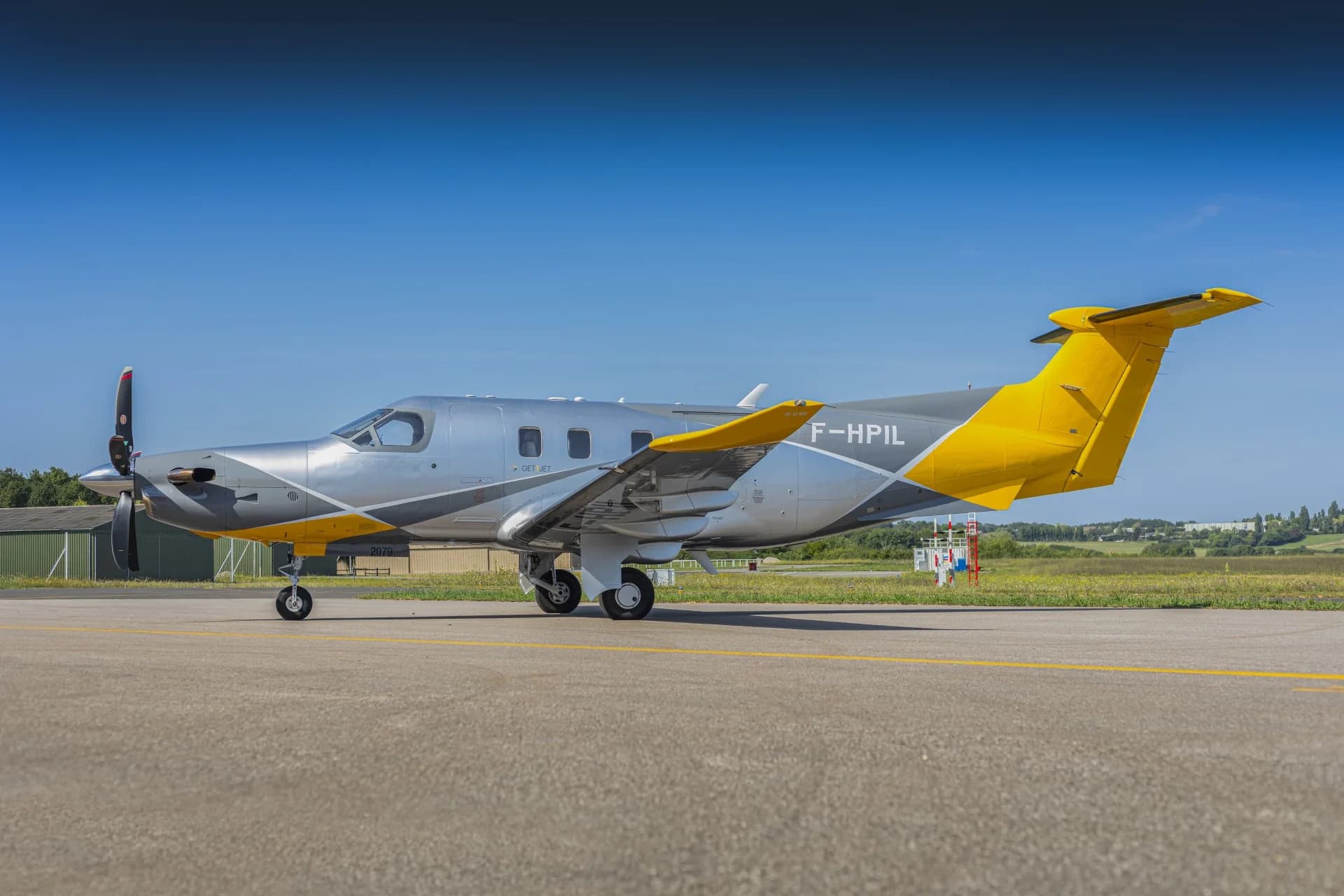 A side view of the silver and yellow Pilatus PC-12 aircraft (F-HPIL) parked on an airfield.