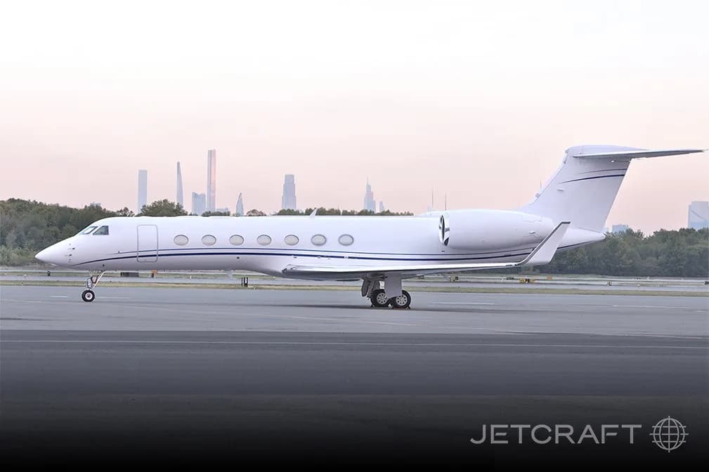 A white Gulfstream G550 business jet with blue stripes on a tarmac with a city skyline in the background at dusk.