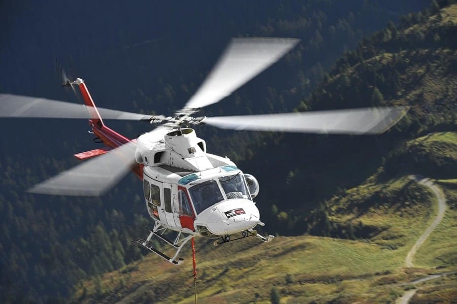 White and red helicopter flying over a mountainous landscape.