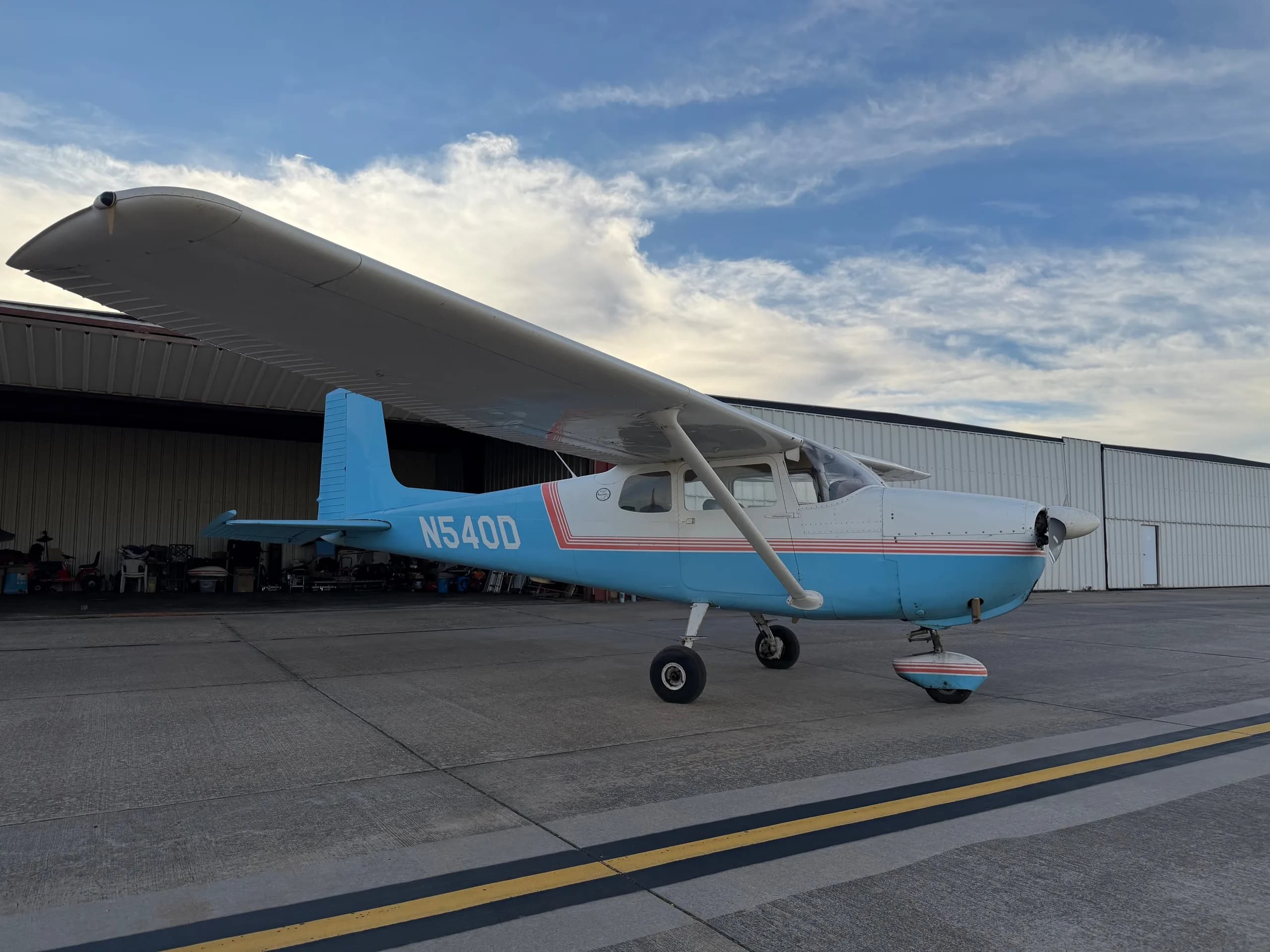A right-side view of a white and light blue Cessna 172, registration N540D, parked on an airfield next to hangars.