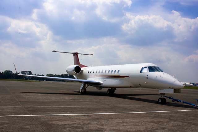 A white business jet with red and gold stripes on the tail and fuselage, parked on an airfield under a cloudy sky.